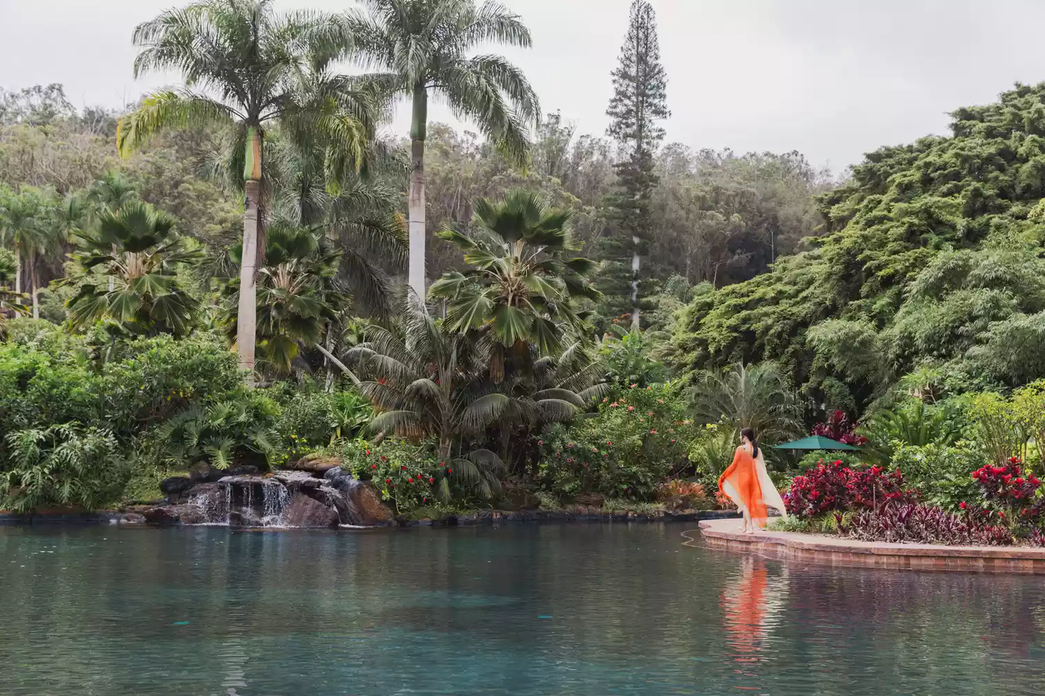 A woman walking along a peaceful, lushly landscaped lagoon at the Sensei Lanai wellness resort.