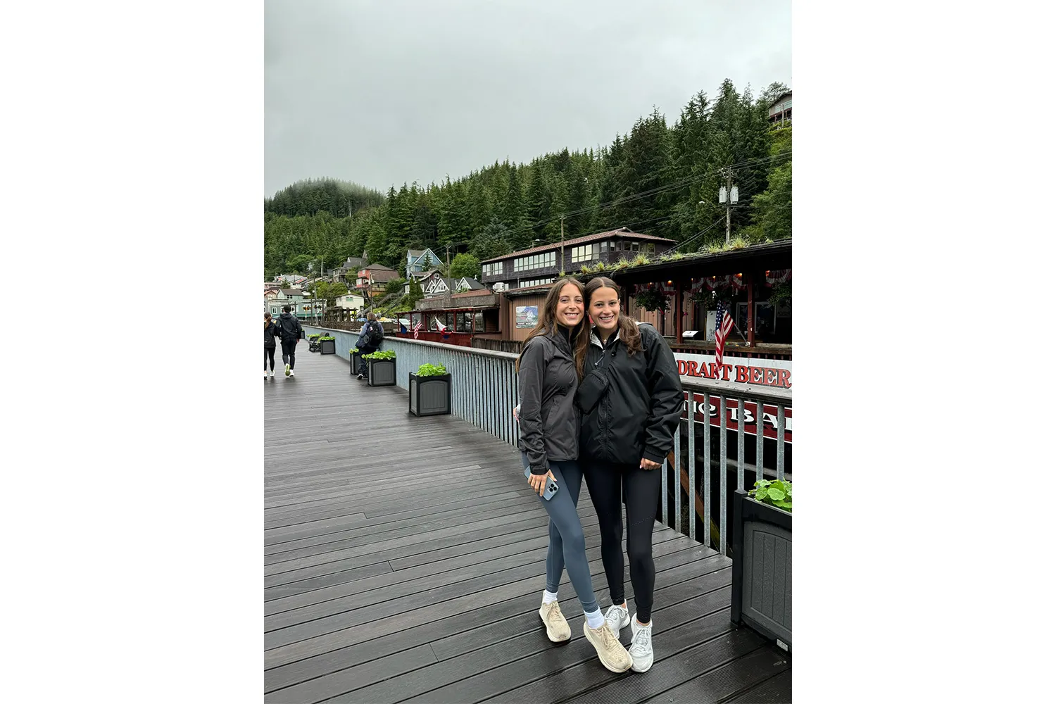 Two people walking on a wooden boardwalk in a forested Alaskan coastal town.