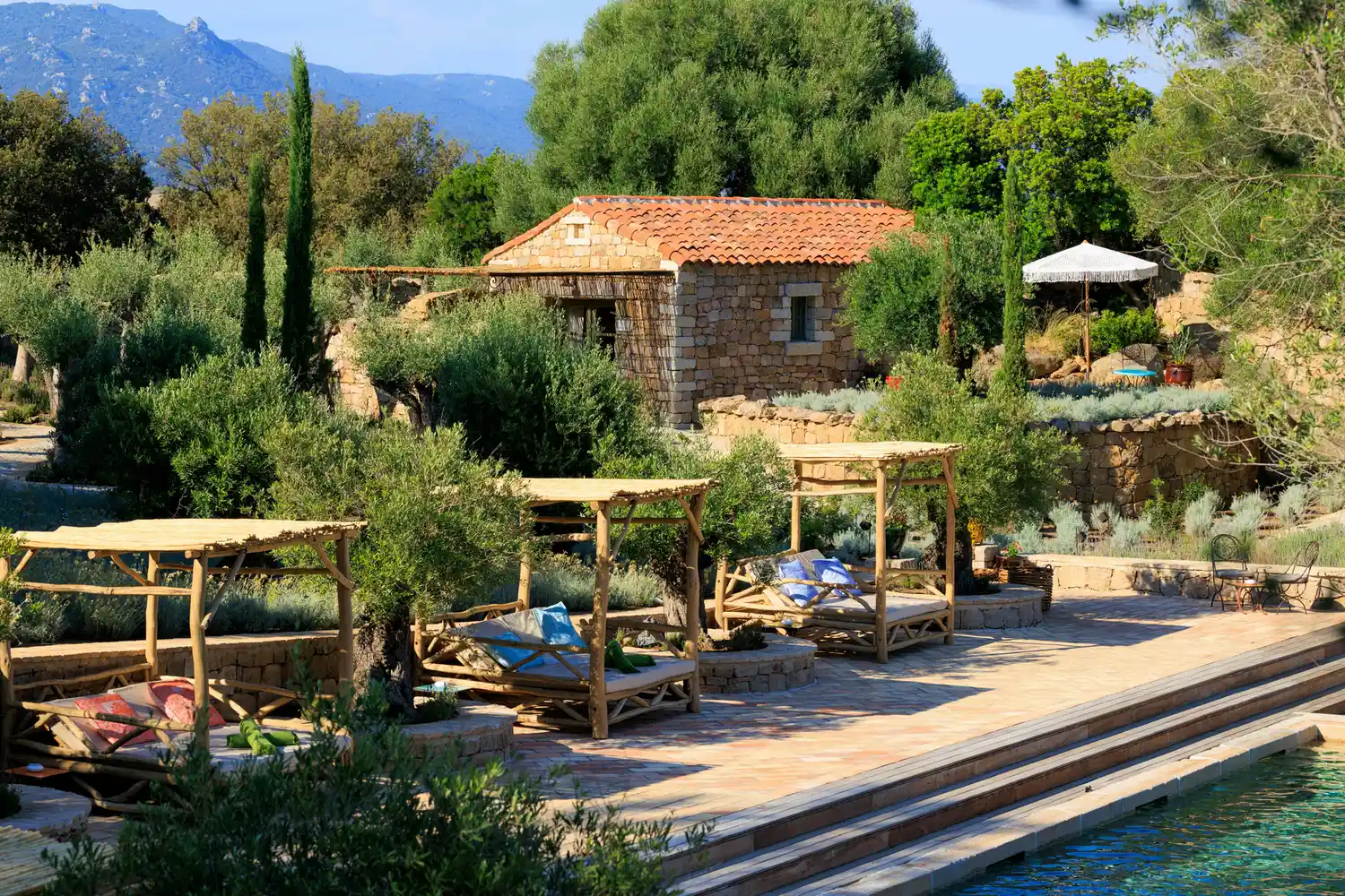 A luxury poolside terrace surrounded by traditional stone cottages and lush Mediterranean greenery.