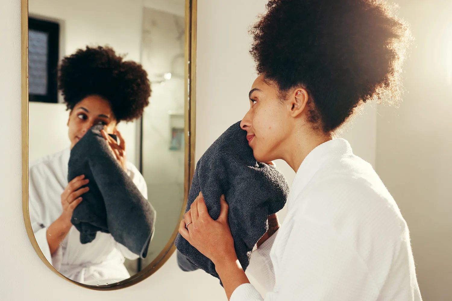 A woman in a bathroom setting using a dark-colored towel to clean her face.