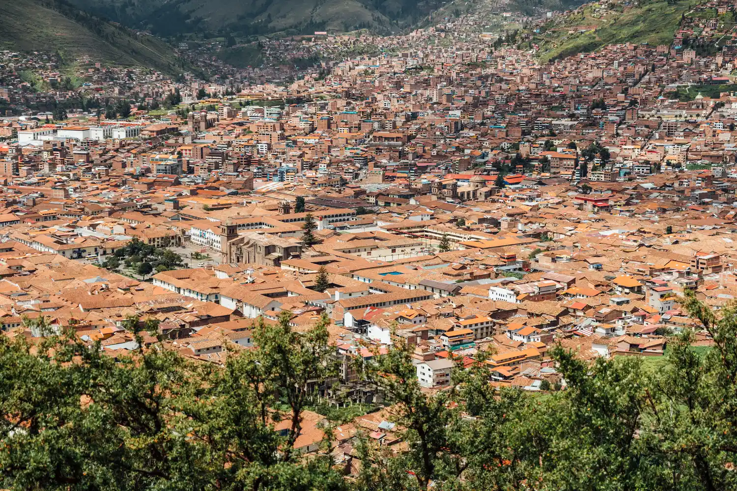 Aerial view of Cuzco, Peru, featuring dense orange-roofed buildings nestled within green mountain peaks.