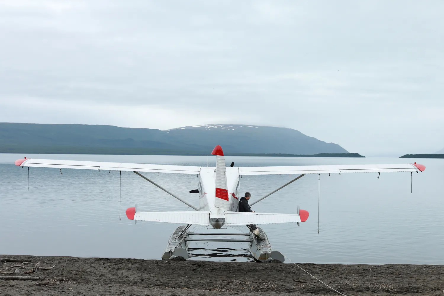 A bush pilot standing by a seaplane on a lake shore with rugged mountains in the distance.