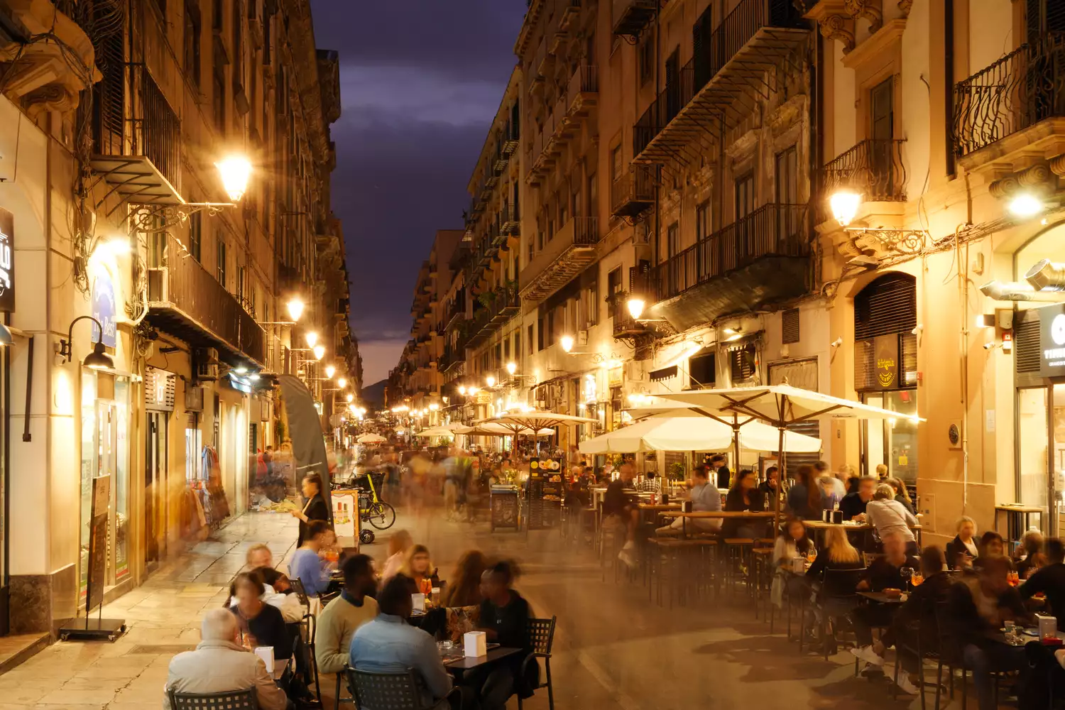 A busy night scene on Via Maqueda in Palermo with people dining at outdoor restaurant tables.