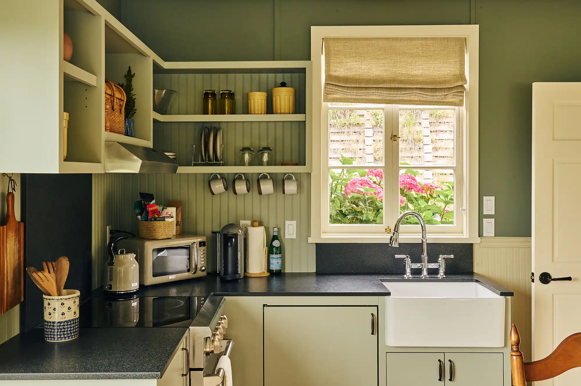 A bright farmhouse kitchen with open white shelving, a deep sink, and a view of spring flowers through the window.