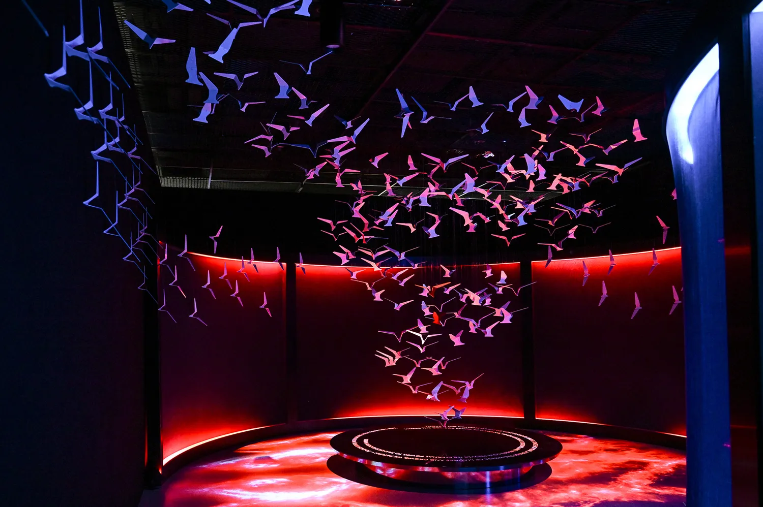 A gallery room bathed in red light featuring suspended glowing geometric shapes and floor reflections.