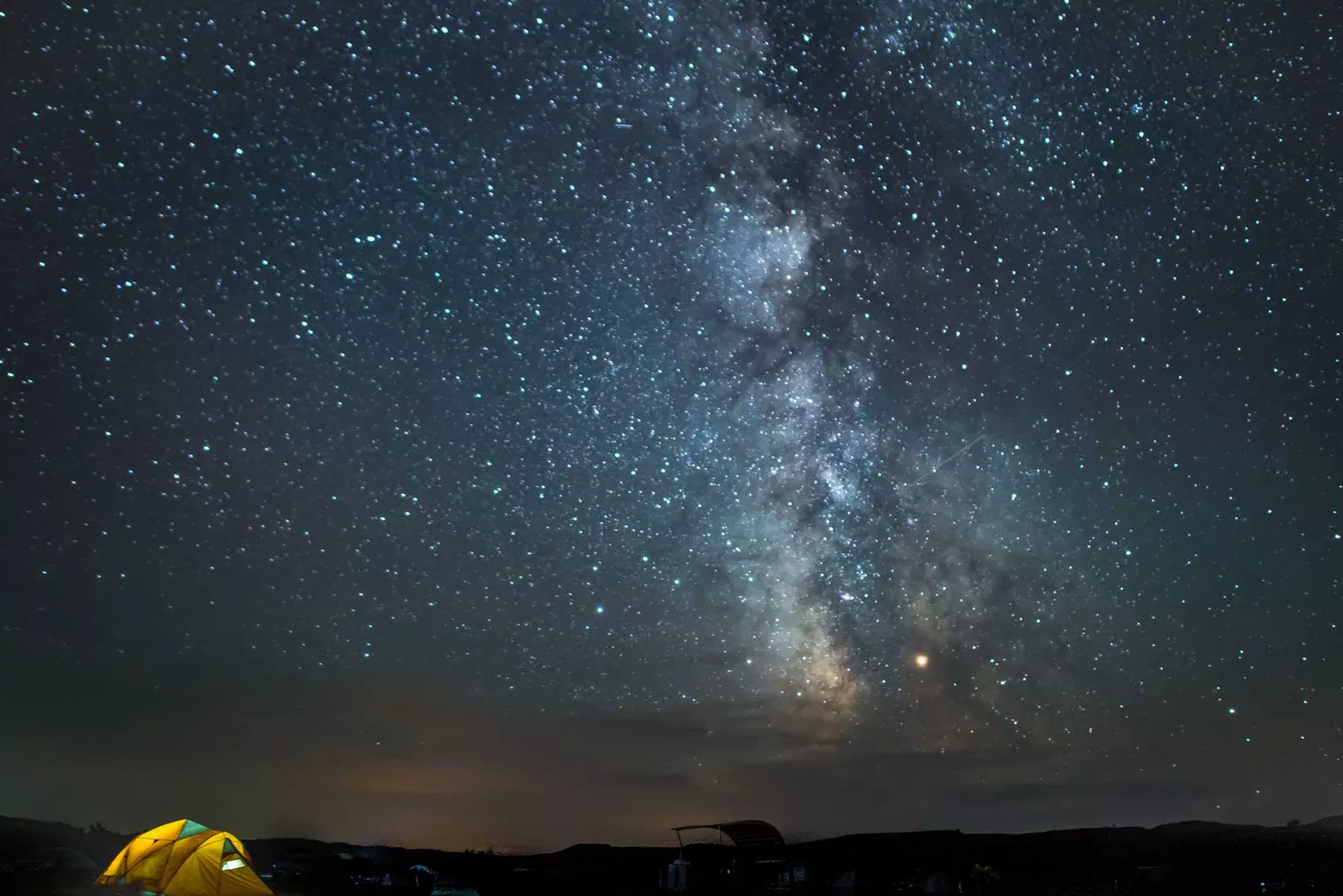 The Milky Way galaxy stretched across a clear dark night sky over a primitive campground setup.