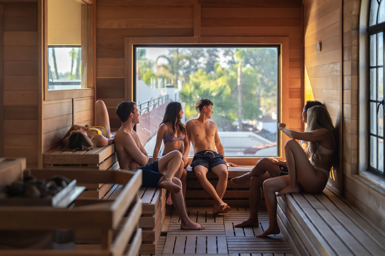 A group of visitors conversing inside a modern wooden sauna with a large scenic window.