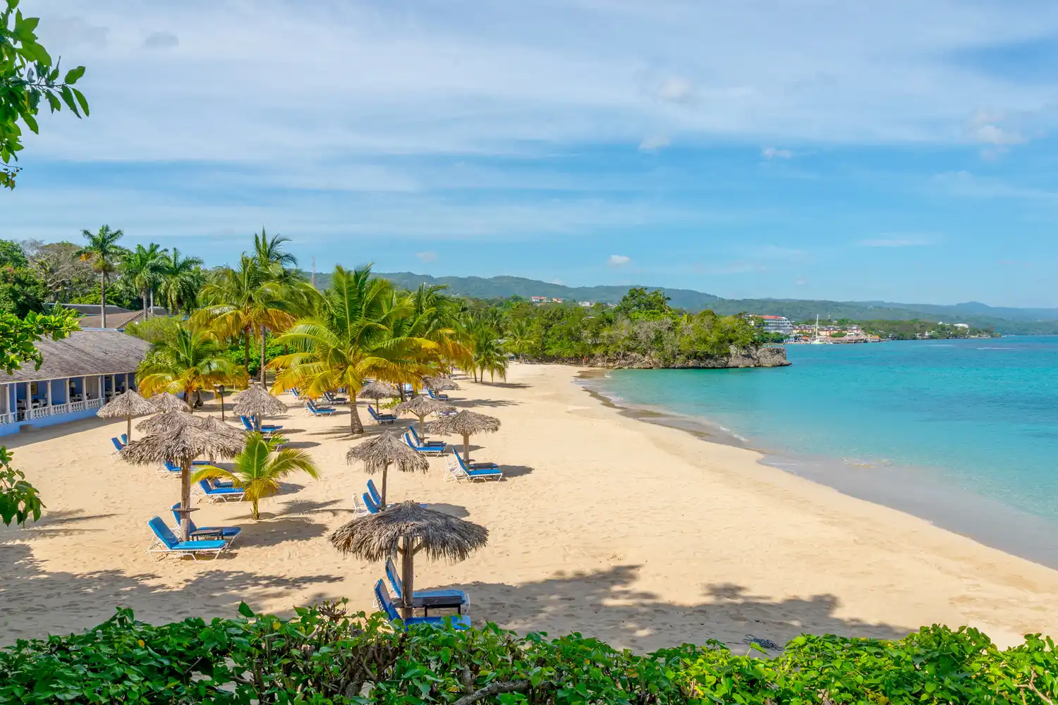 A peaceful white sand beach at Jamaica Inn with blue umbrellas, lounge chairs, and palm trees.