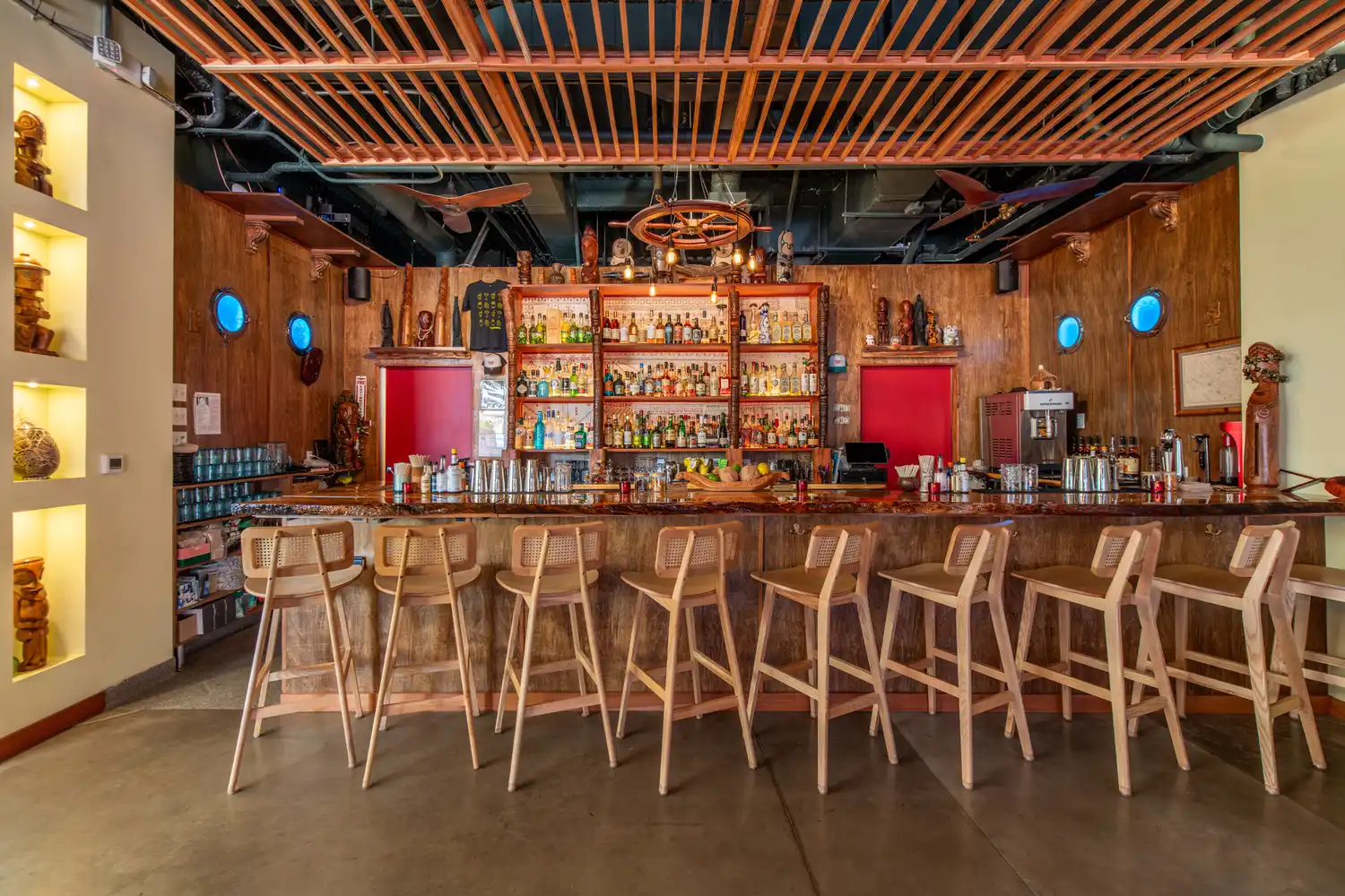 A wooden bar with stools and shelves stocked with various spirits under a wooden slat ceiling at Tikehau Lounge.