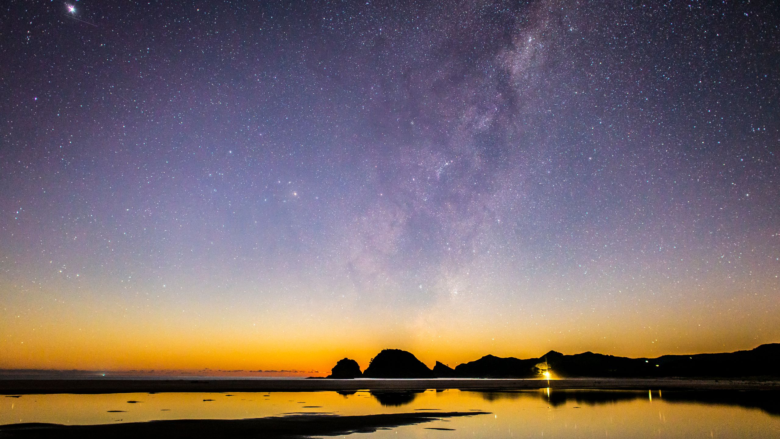 The starry night sky reflecting over the waves and shore of a beach on Great Barrier Island.
