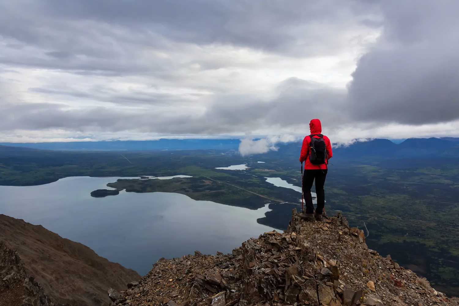 A hiker at the edge of a rocky cliff overlooking a deep blue lake surrounded by mountains.