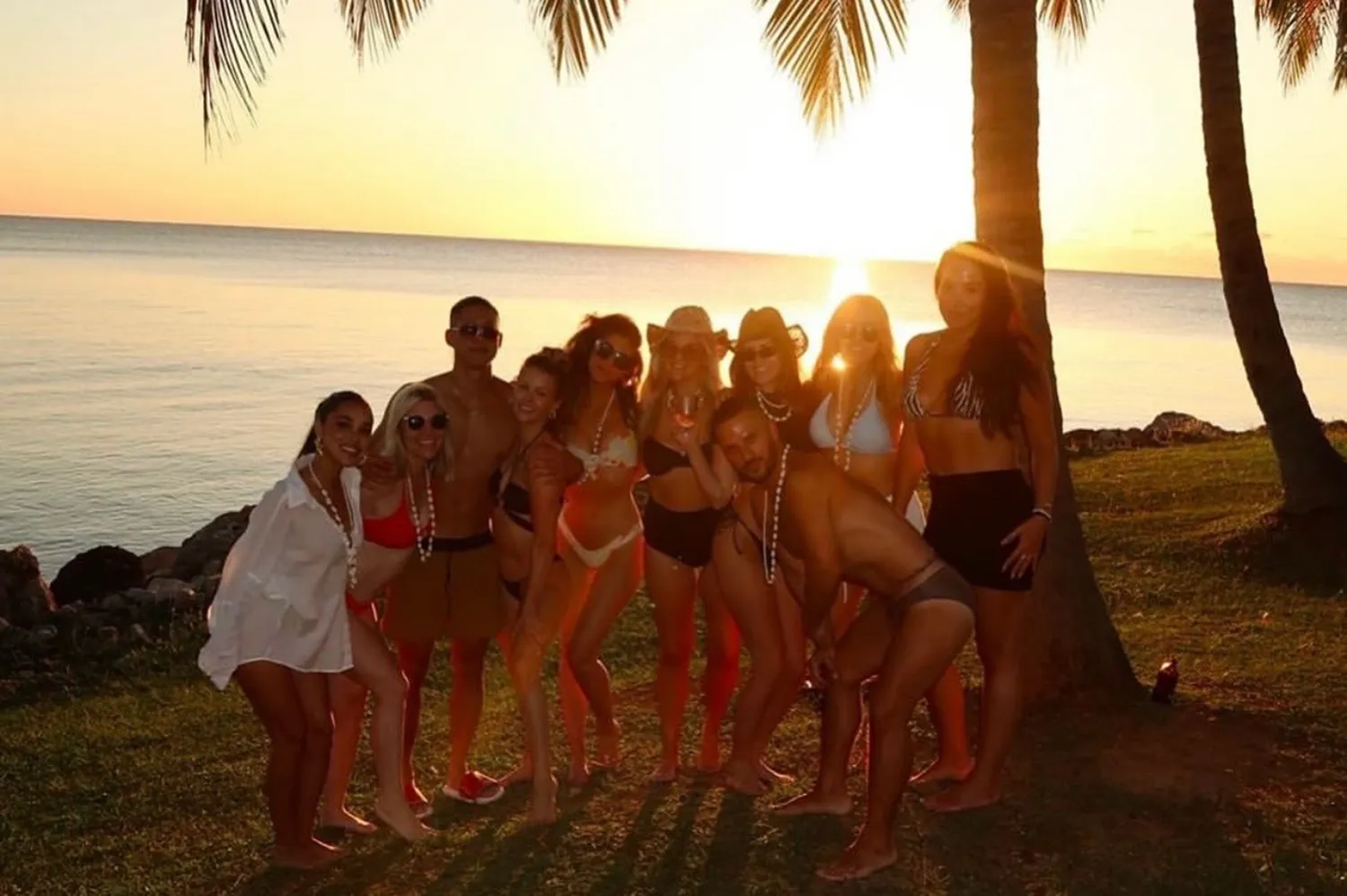 A group of people standing together by the water during a gold-tinted sunset near palm trees.