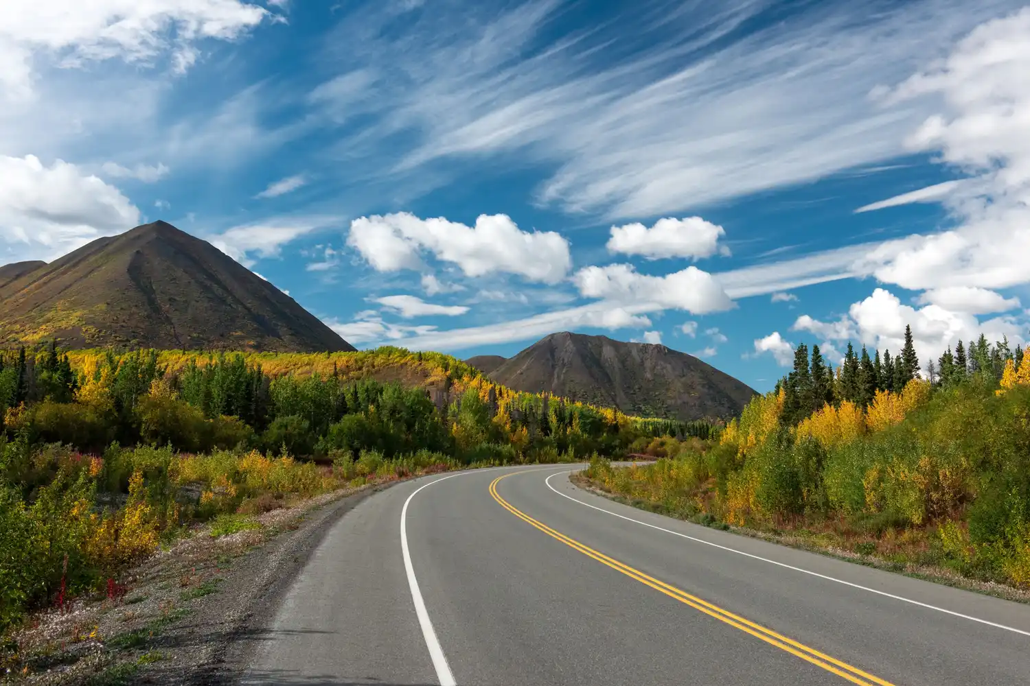A long, winding asphalt road leading toward distant, rugged mountains through a forested landscape.
