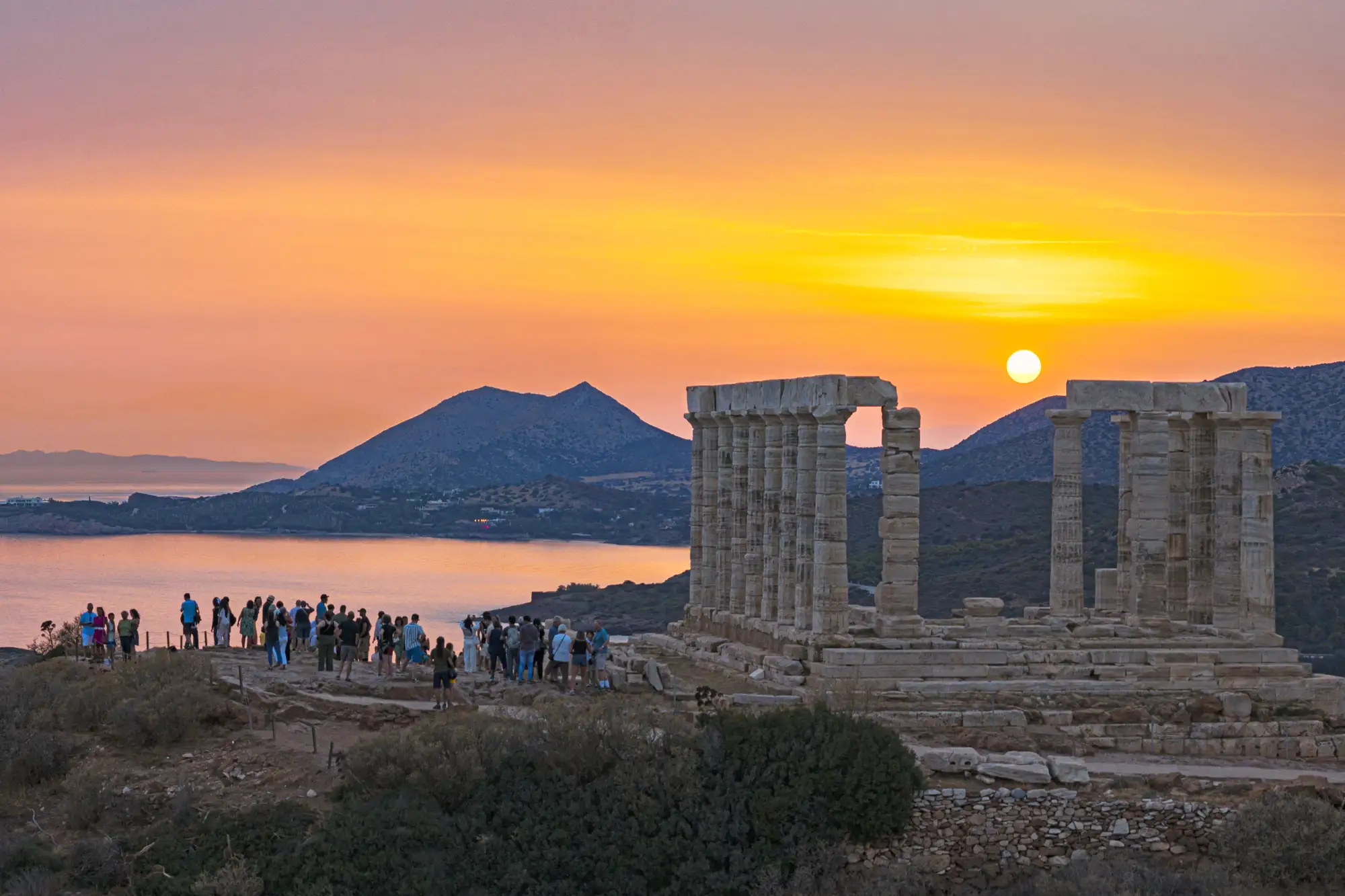 The ancient Temple of Poseidon at Cape Sounion silhouetted against a vibrant orange and pink sunset sky.