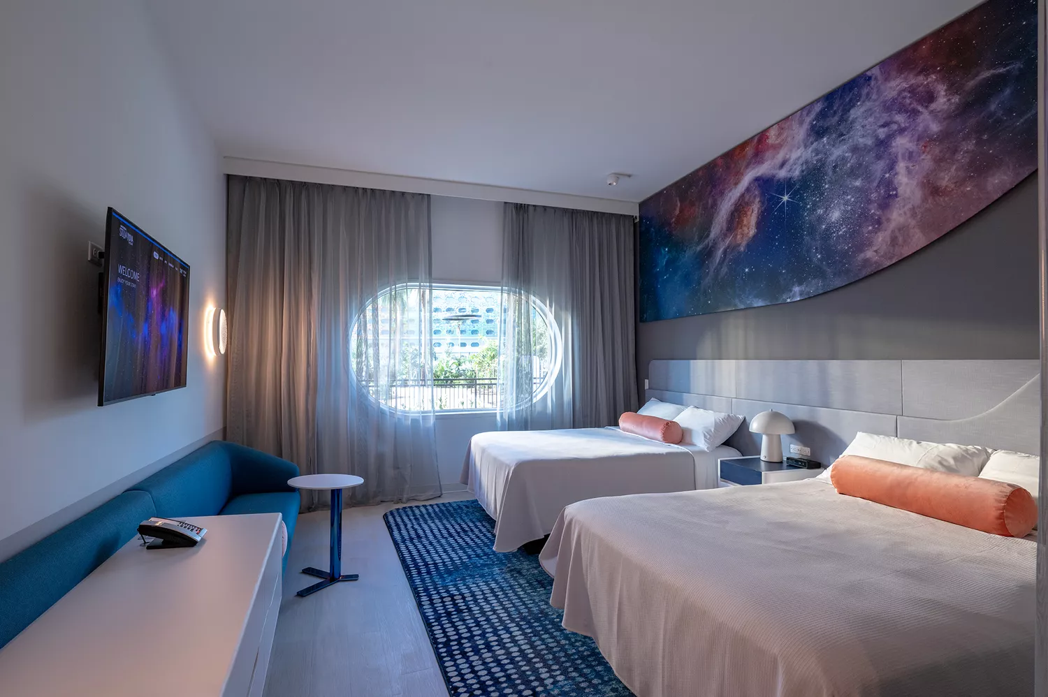 A standard guest room at Universal Stella Nova Resort with two queen beds and a circular porthole window.