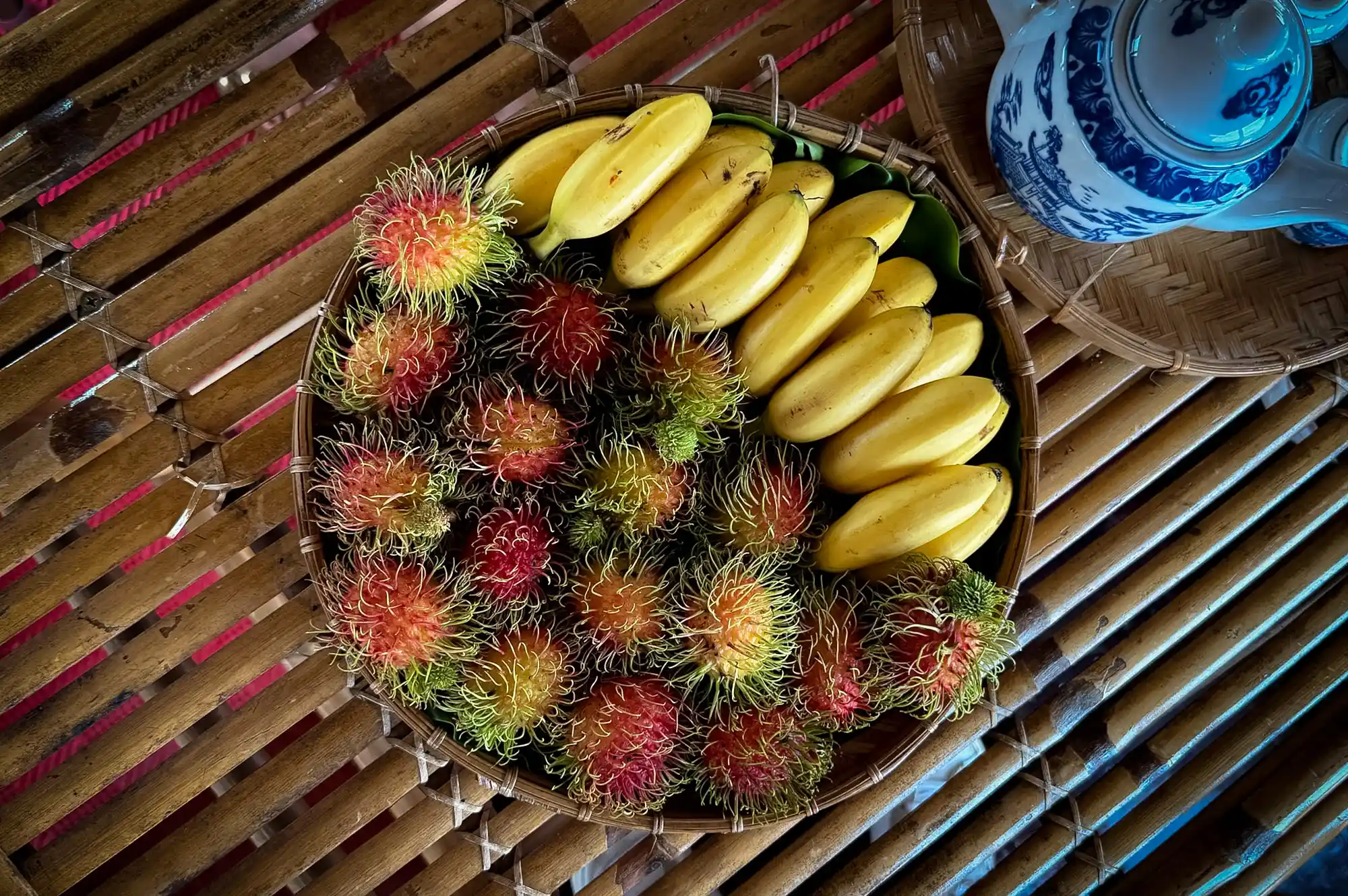 A basket of tropical fruits including bananas and rambutan on a bamboo surface.