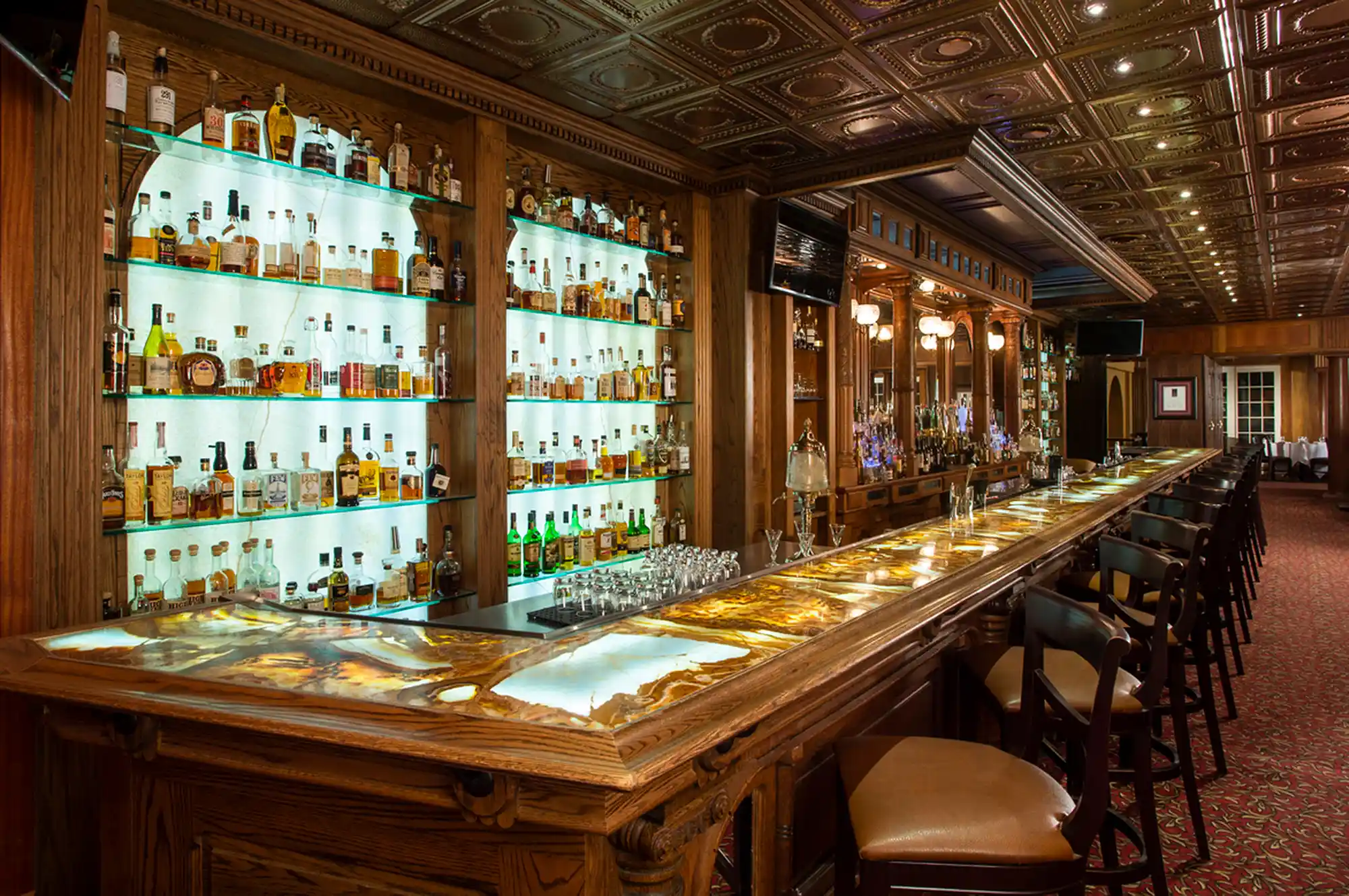 Elegant interior of a hotel bar with glowing shelves filled with diverse whiskey bottles and dark wood seating.