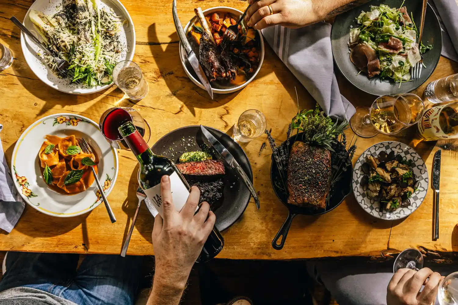 An overhead view of a dinner table filled with gourmet dishes and a person pouring a glass of wine.