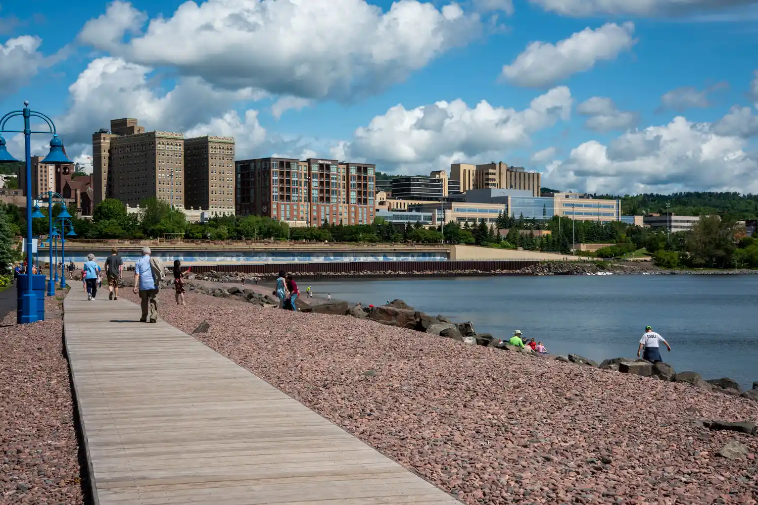 Pedestrians walking along the paved Lakewalk path with the rocky Lake Superior shoreline in view.