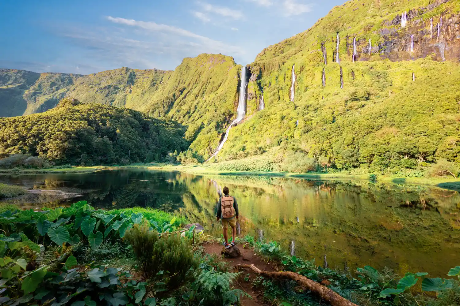 A person standing before a massive green cliff with multiple waterfalls in the Azores.