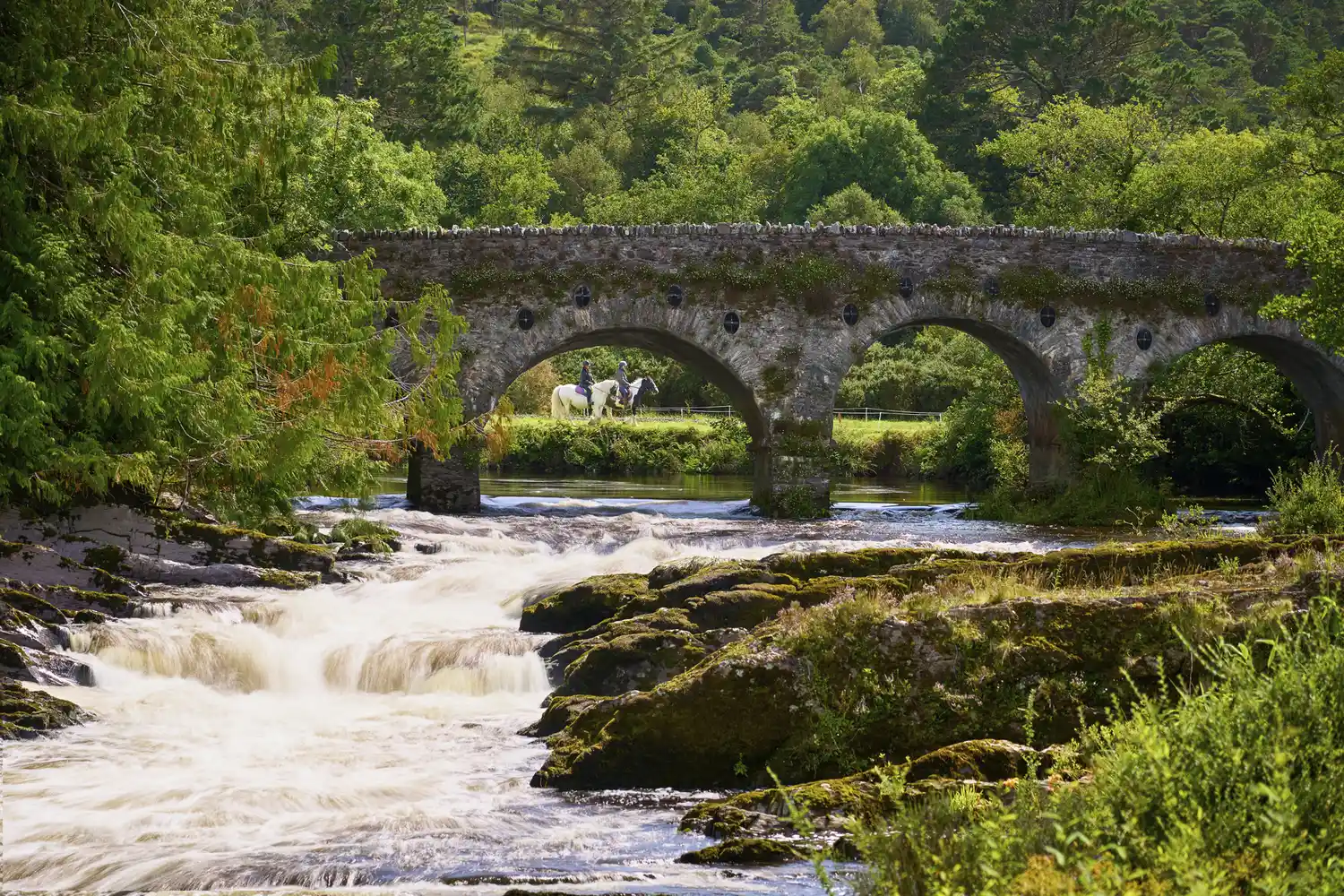A person riding a horse across an old stone bridge over a flowing mountain stream.