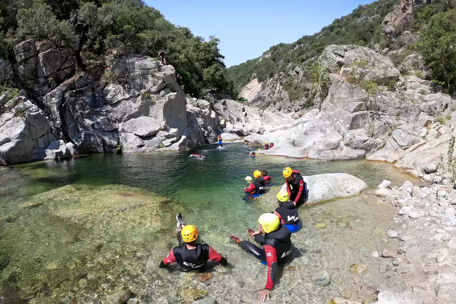 A group of adventurers wearing wetsuits and helmets resting in the clear, rocky pools of the Travo River.