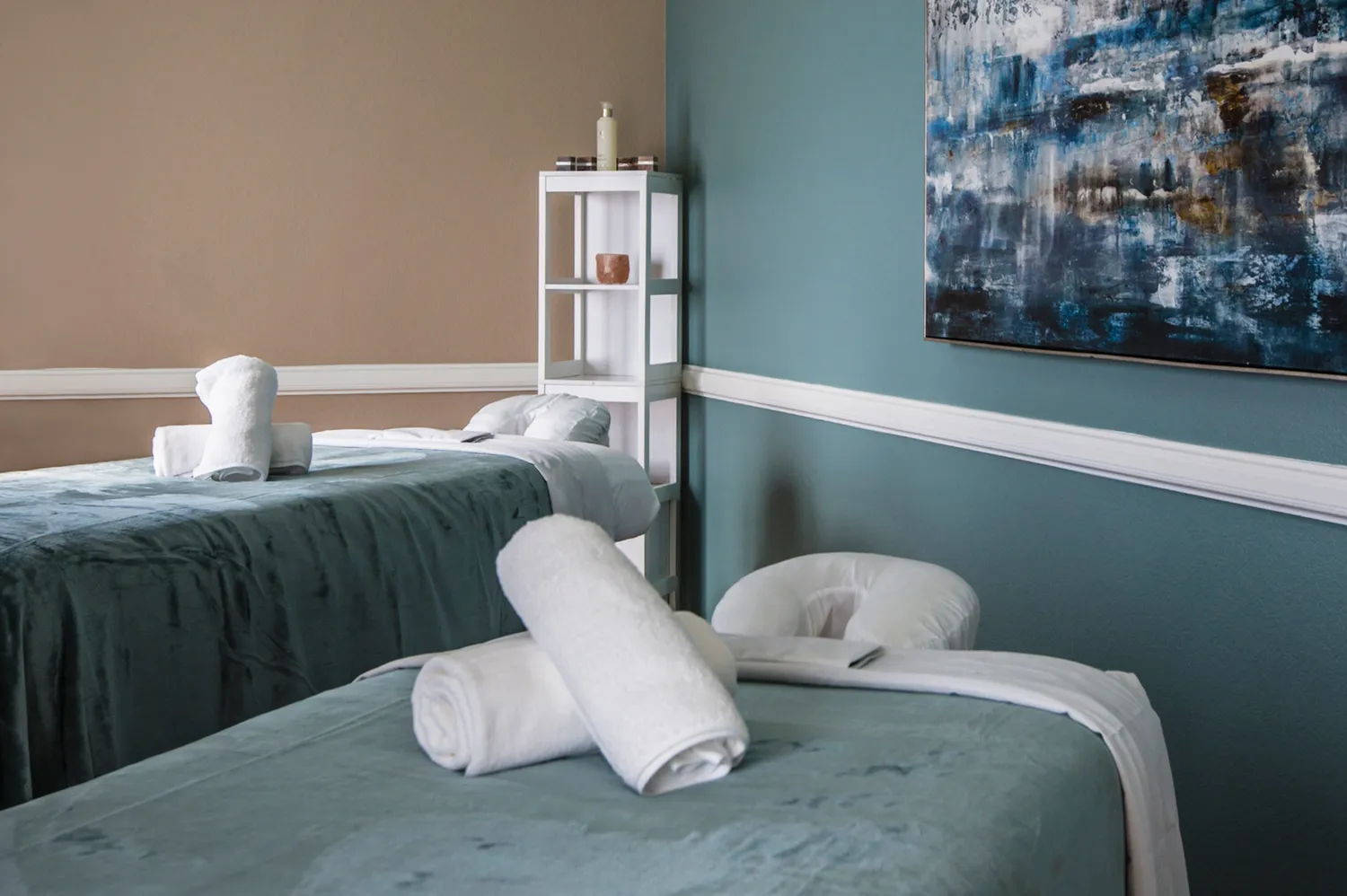 Two modern spa treatment tables prepared with rolled towels in a serene room with teal and beige walls.