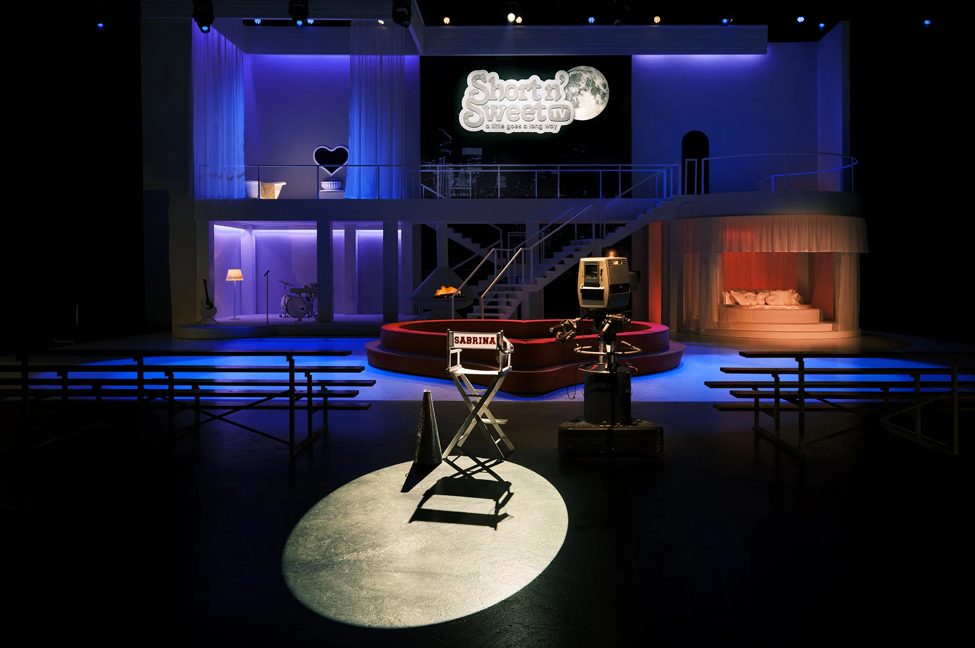A theater-style stage set with director chairs, stage lighting, and cozy bed arrangements.