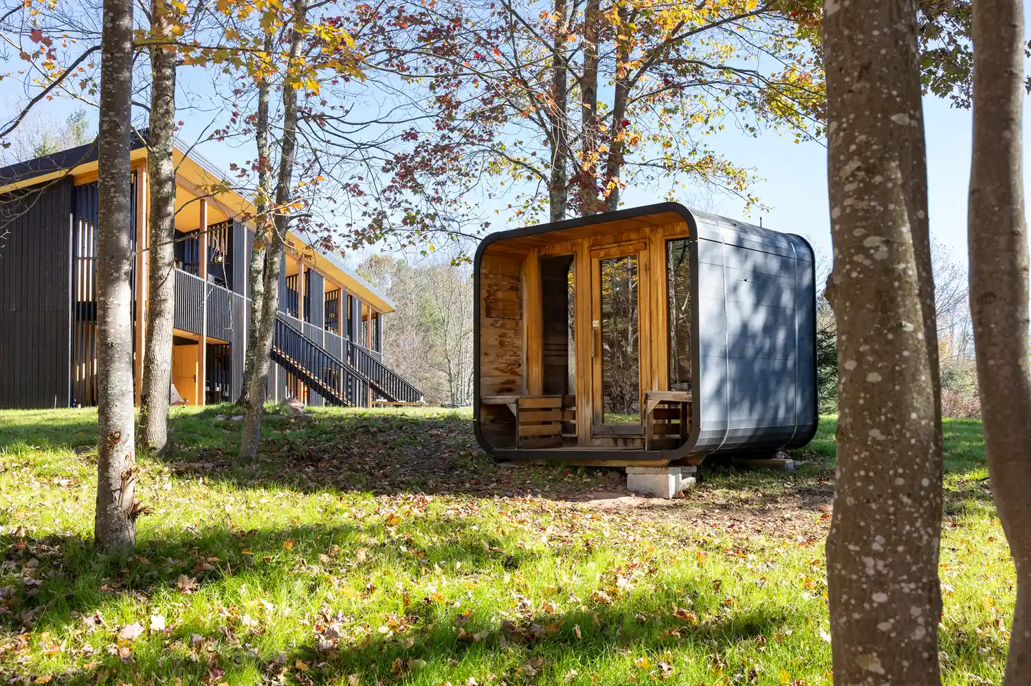 A standalone wood-barrel sauna located near the hotel house within a wooded area.