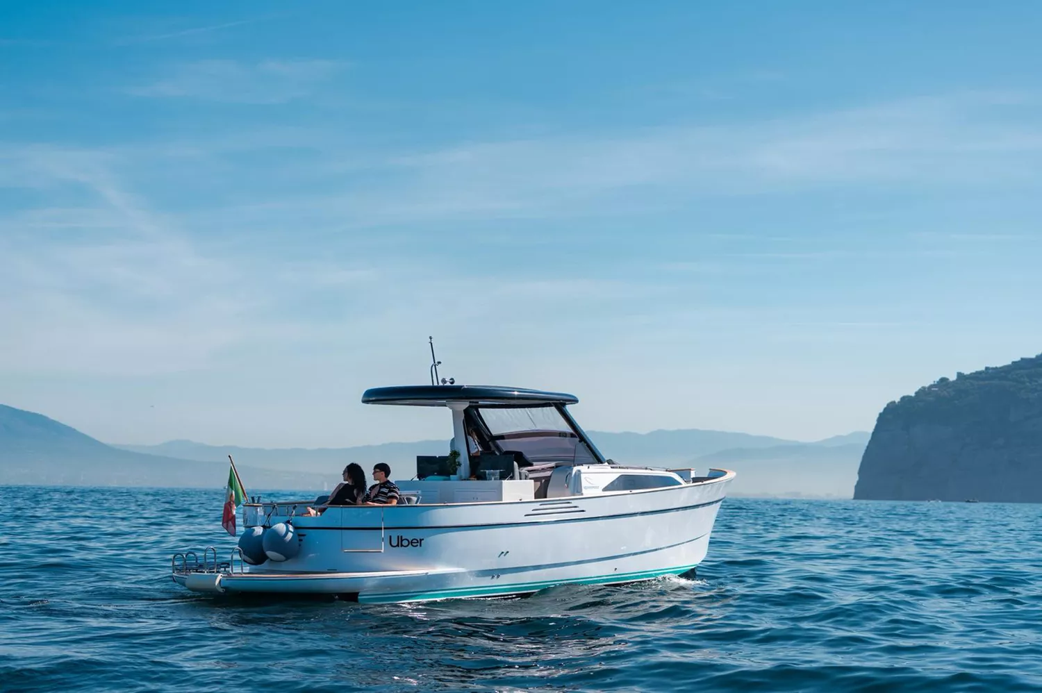 A small motorboat with people on board floating on calm blue water with hills in the background.