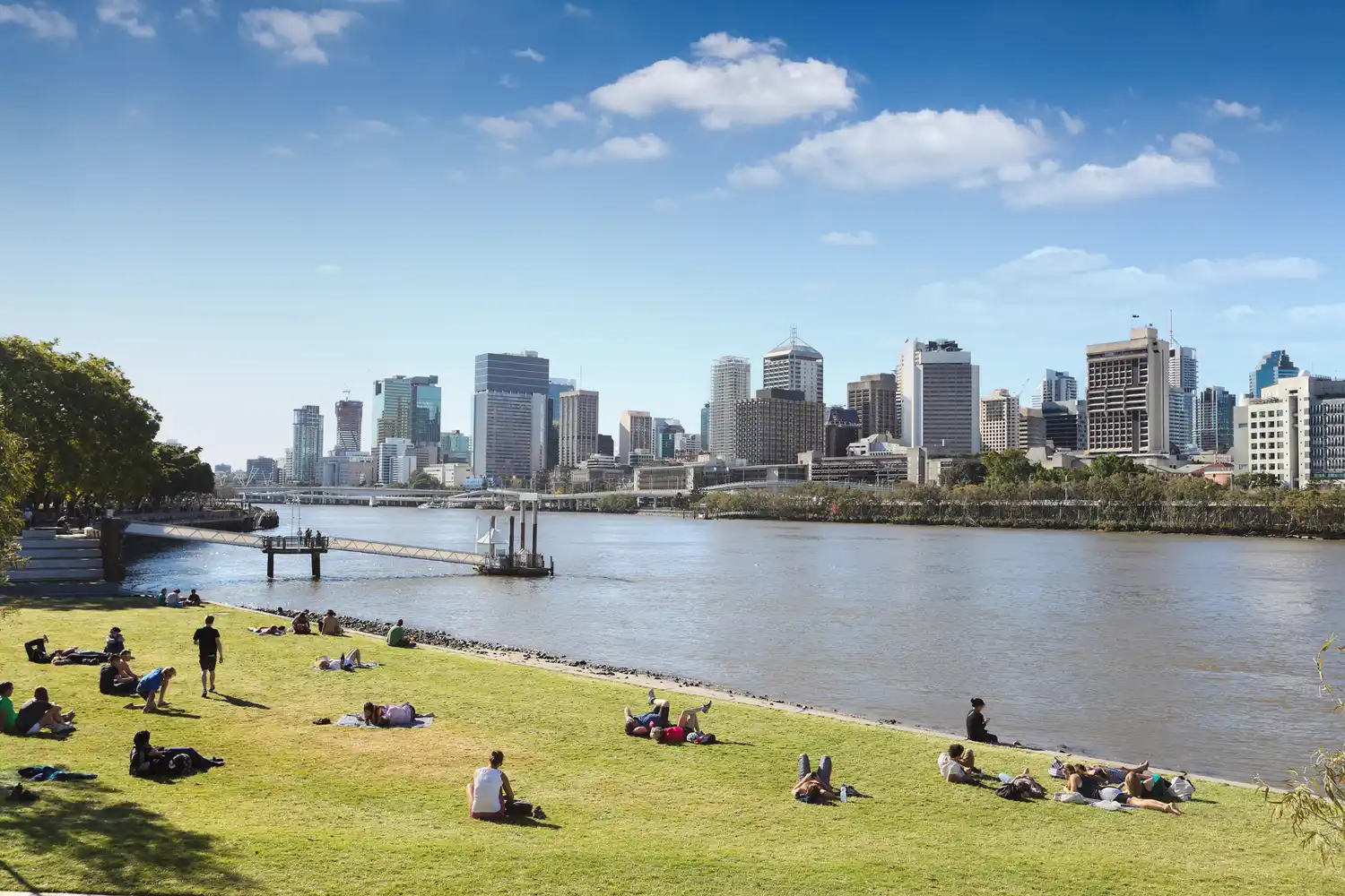 Families relaxing on a lush lawn at South Bank with the Brisbane city skyline across the river.