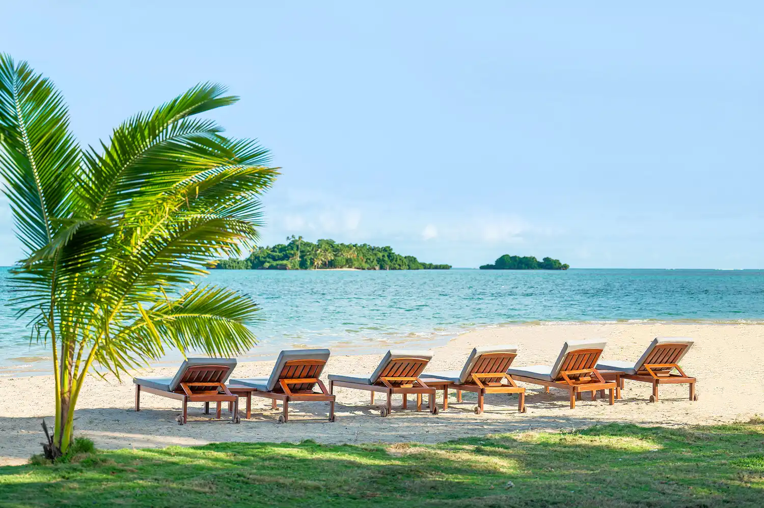 Peaceful scene of lounge chairs on a sandy beach with palm trees and a view of distant islands.