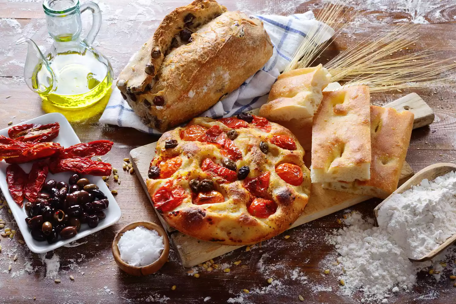 Freshly baked Italian breads and Mediterranean ingredients on a floured wooden work surface.