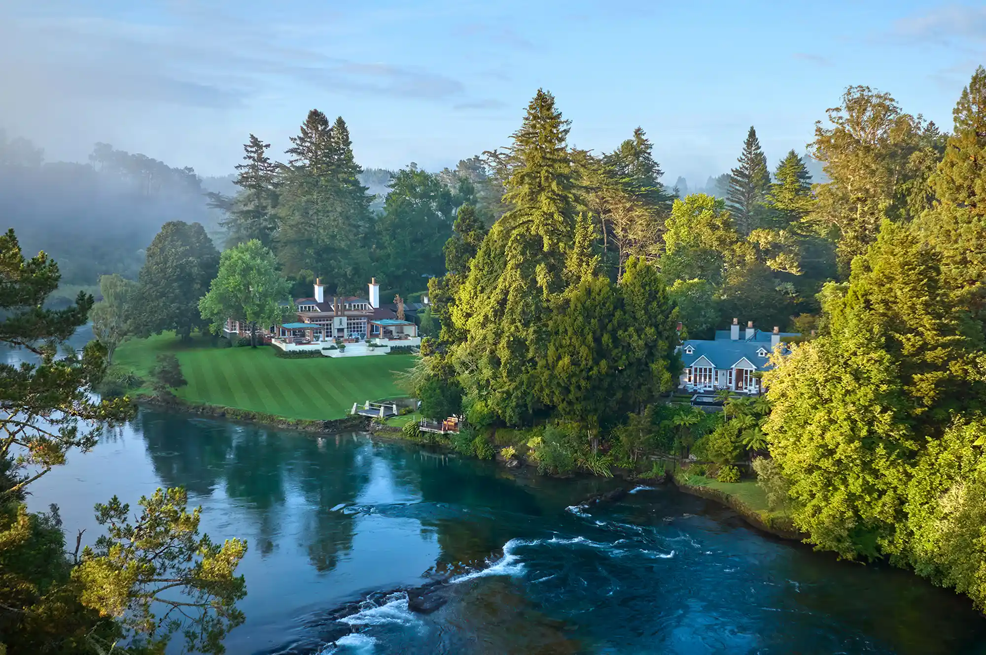 An estate-style lodge with green gardens situated prominently next to a flowing river.
