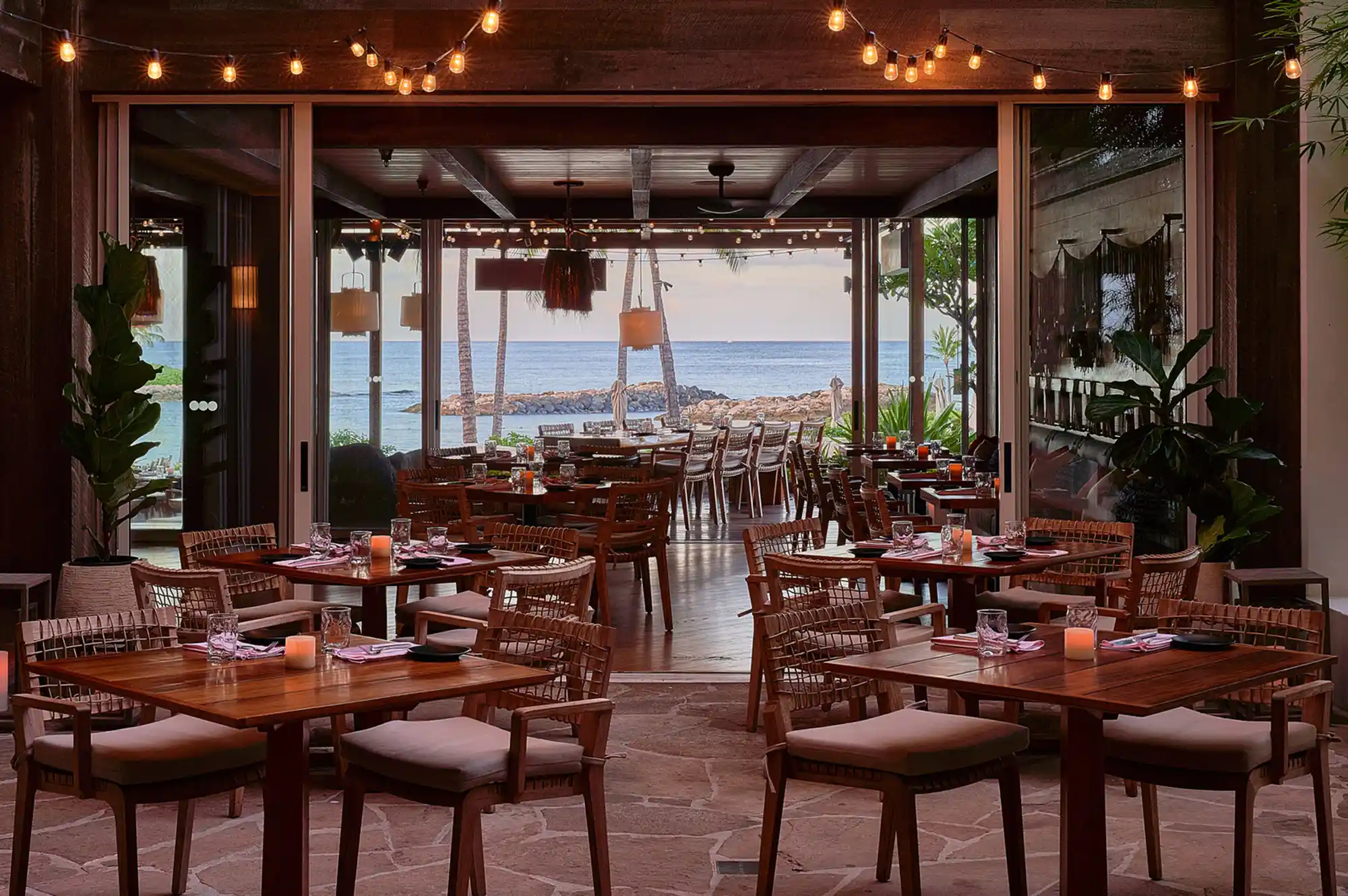 Outdoor dining area with string lights and open windows overlooking the ocean at sunset.