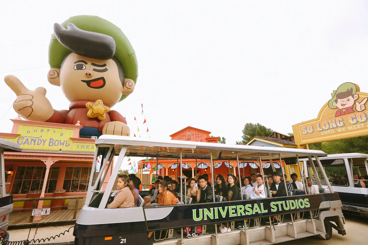 A multi-car studio tour tram passing by large character displays on the backlot.
