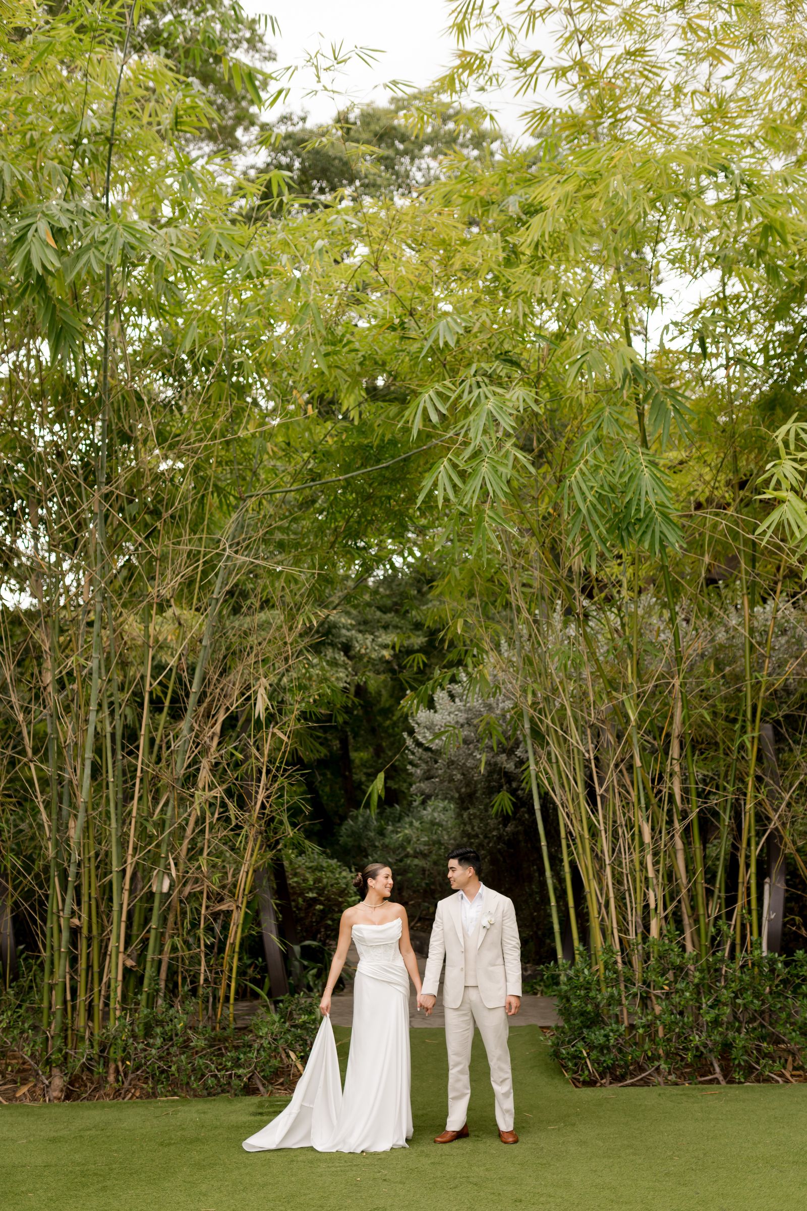 A bride in a white gown standing in a lush tropical grove.